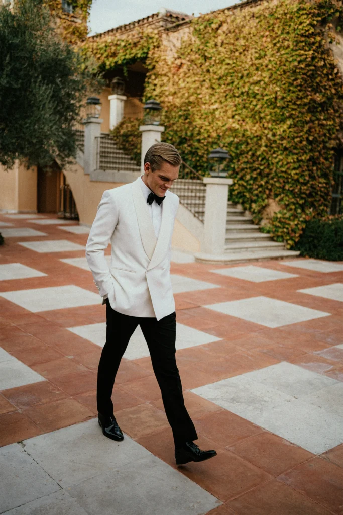 A man in a white dinner jacket, black trousers, and black shoes walks outdoors on a tiled patio near a building covered in ivy.