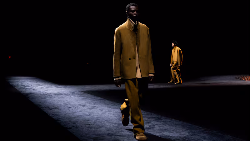 A male model walks down a dimly lit catwalk wearing mustard yellow trousers and a matching jacket, capturing the emotion and desire often sought in luxury retail; another model is visible in the background.