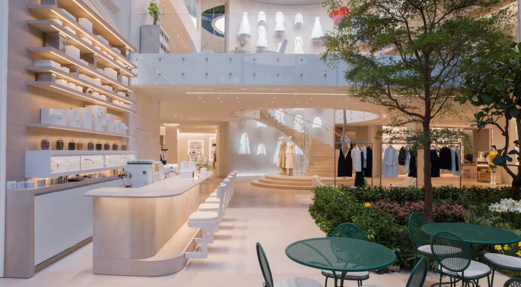 A modern luxury retail store with a minimalist café, spiral staircase, and curated clothing displays evokes desire and emotion. Green patio furniture and indoor plants complement the neutral-toned design in the inviting foreground.