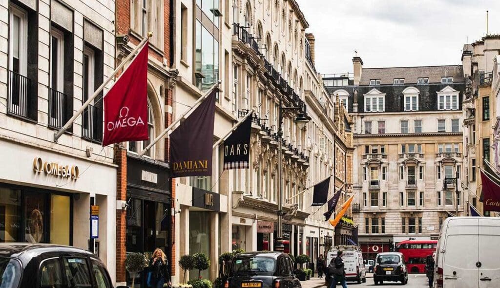 A city street exuding desire, lined with luxury retail shops like Omega and Damiani, vibrant flags fluttering above, as pedestrians and parked cars mingle with a red double-decker bus in the background.